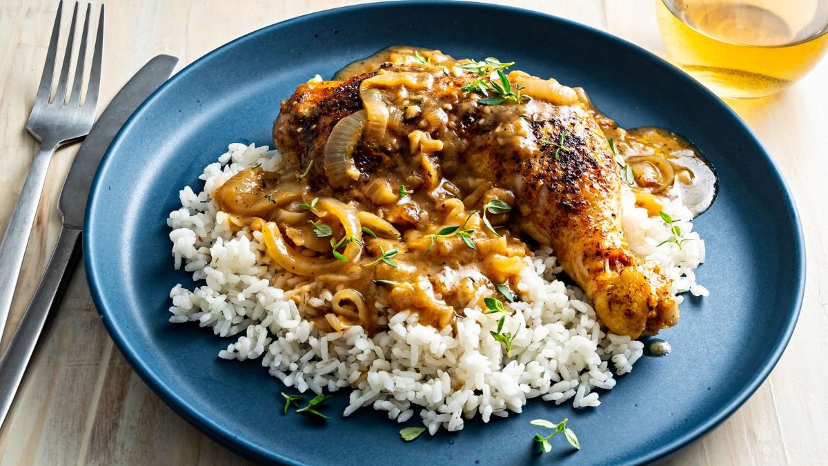 smothered chicken served over rice on a black plate with a glass of apple juice