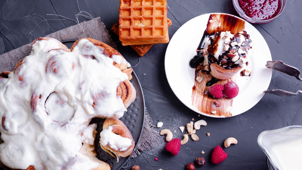 a Rhodes Cinnamon Roll with creamy icing, and topped with chocolate sauce, crushed cashews, and raspberry jam, served on a plate next to an oval pan full of freshly baked cinnamon rolls